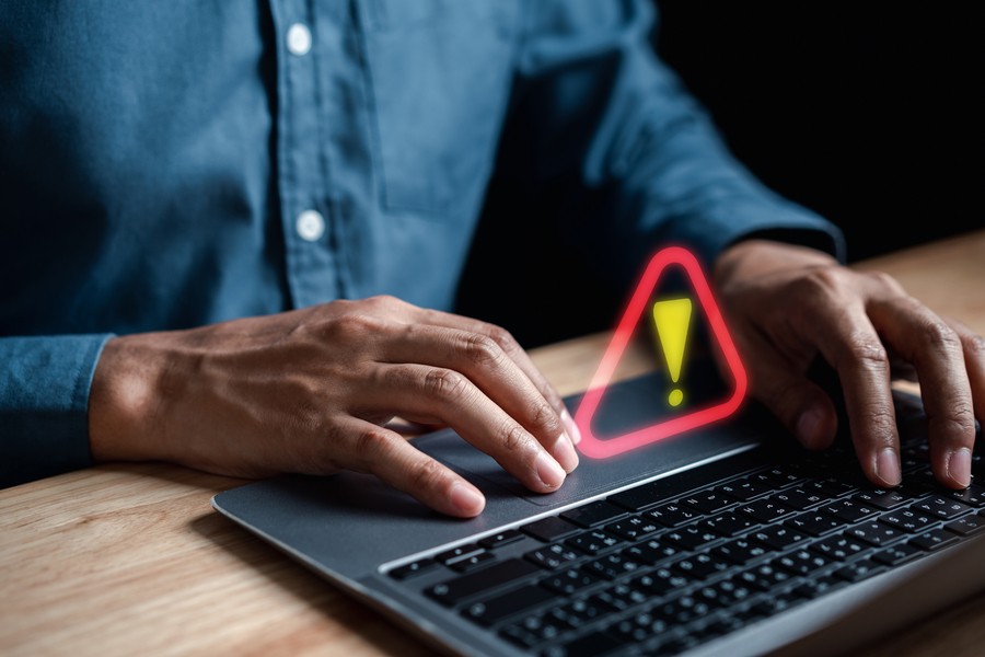 A man’s hands typing on a laptop keyboard, with a glowing red warning sign appearing to float above the keyboard, suggesting computer error.