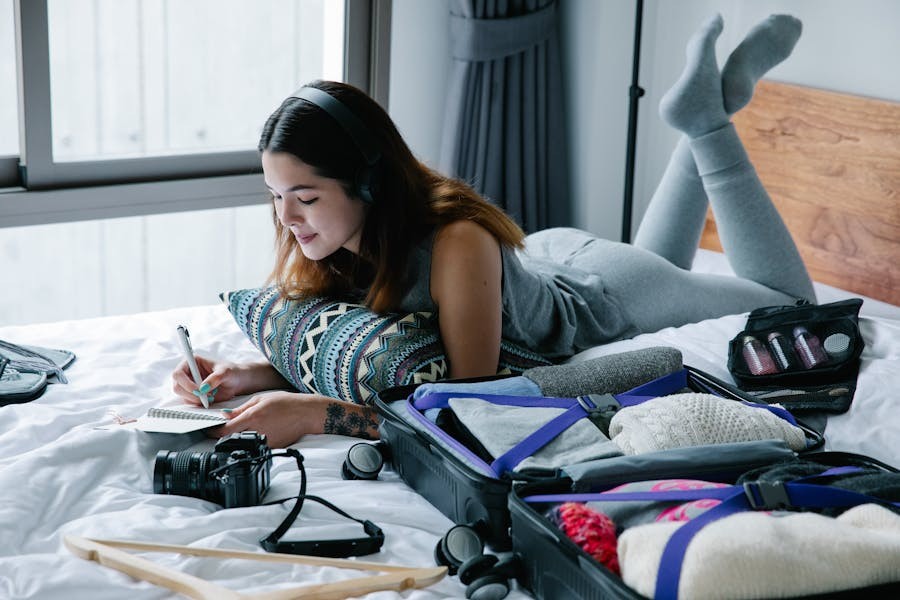 Woman lying on bed packing a suitcase, writing in a notebook with headphones on, preparing for travel.